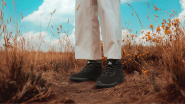 Person wearing loose white trousers and black boots standing in a dry grass field under a blue sky with white clouds