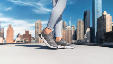 Close-up of grey Giesswein Merino Wool trainers on the feet of a runner wearing light blue leggings on a rooftop terrace with high-rise buildings in the background.
