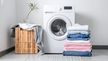 A white front-loading washing machine in a bright bathroom. To the left of it is a wooden box covered with a grey cloth; to the right is a neatly stacked pile of laundry. In the background is a green houseplant.
