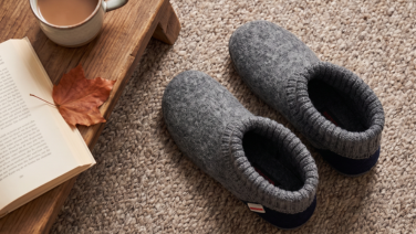 A top view of a pair of grey merino wool slippers with a knitted upper and a dark blue sole on a beige carpet; next to them, an open book, a maple leaf and a coffee cup on a wooden tray.