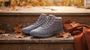 A pair of grey merino wool boots with a high shaft, laces and a label stand on rustic wooden floorboards, framed by autumn leaves and a rust-red blanket in the warm light.