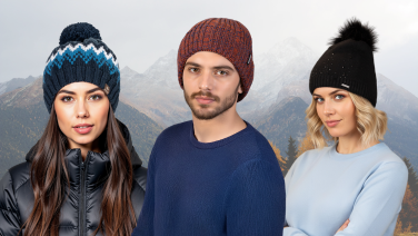 Three young people wearing colourful knitted hats in front of a snowy mountain landscape