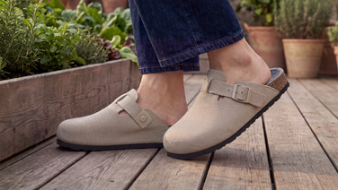 A close-up of a person’s feet in dark blue jeans, standing on a wooden decking. They are wearing beige merino clogs with buckled straps and cork soles