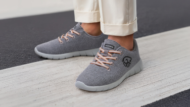 Close-up of a person’s feet in cream-colored, rolled-up pants. They’re wearing gray Merino wool sneakers with apricot-colored laces and a light gray sole. The person is standing on a crosswalk.