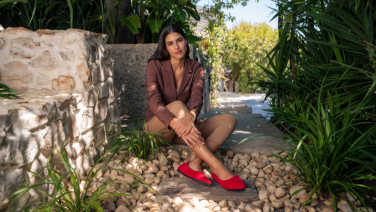 A woman in a brown suit is sitting in front of a stone wall in a Mediterranean garden; she is wearing red Giesswein ballerinas
