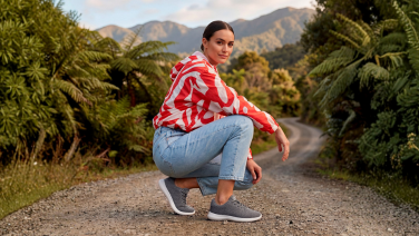 A woman wearing a patterned top and light-coloured jeans is crouching on a gravel path, surrounded by green ferns, with mountains in the background. She is wearing grey merino wool trainers.
