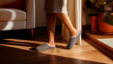 Person in beige clothing wearing light grey Giesswein wool slippers on wooden floor in sun-filled living room