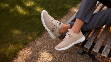 Close-up of beige Giesswein merino sneakers with white sole and embossed logo on the feet of a person in gray pants on a park bench.