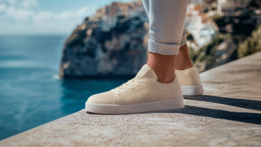 Close-up of a cream-colored Giesswein Merino sneaker with a white flat sole on a stone wall against a Mediterranean seascape.