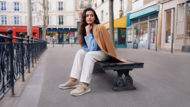 Woman sitting on a municipal bench in a square in the city centre, wearing a beige jacket, blue shirt, white trousers and beige Giesswein Merino Runners trainers.