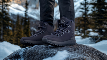 Person wearing dark hiking boots standing on snow-covered rock, winter forest in blurred background